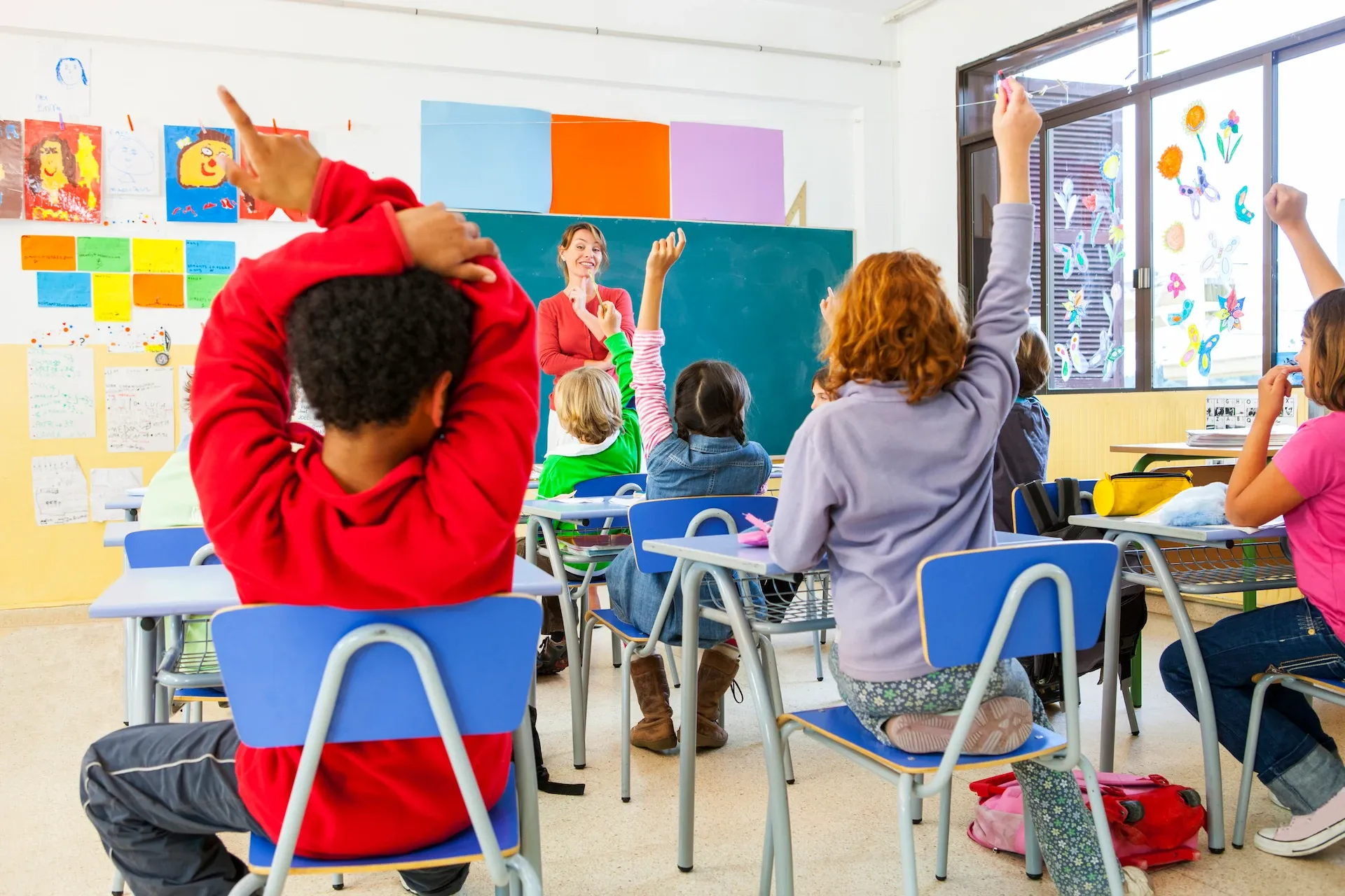 Children at classroom desks raising their hands as a teacher calls on them