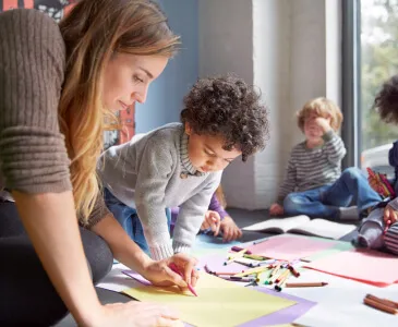 A teacher kneeling beside children drawing on the classroom floor