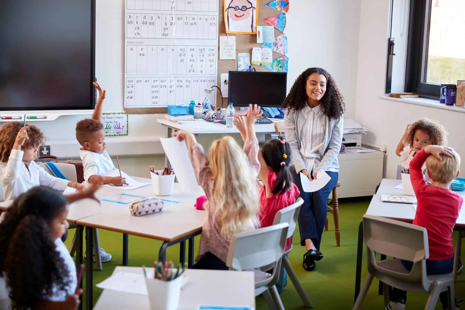 A teacher smiling as children raise their hands at classroom tables