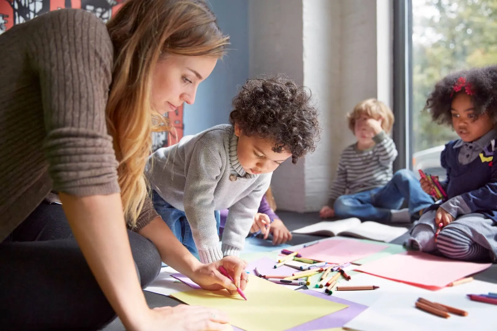 A teacher drawing with a young boy while other children color on the classroom floor